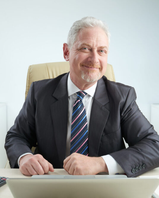 Bearded elderly entrepreneur in formalwear sitting at workplace and looking at camera with wide smile, office interior on background